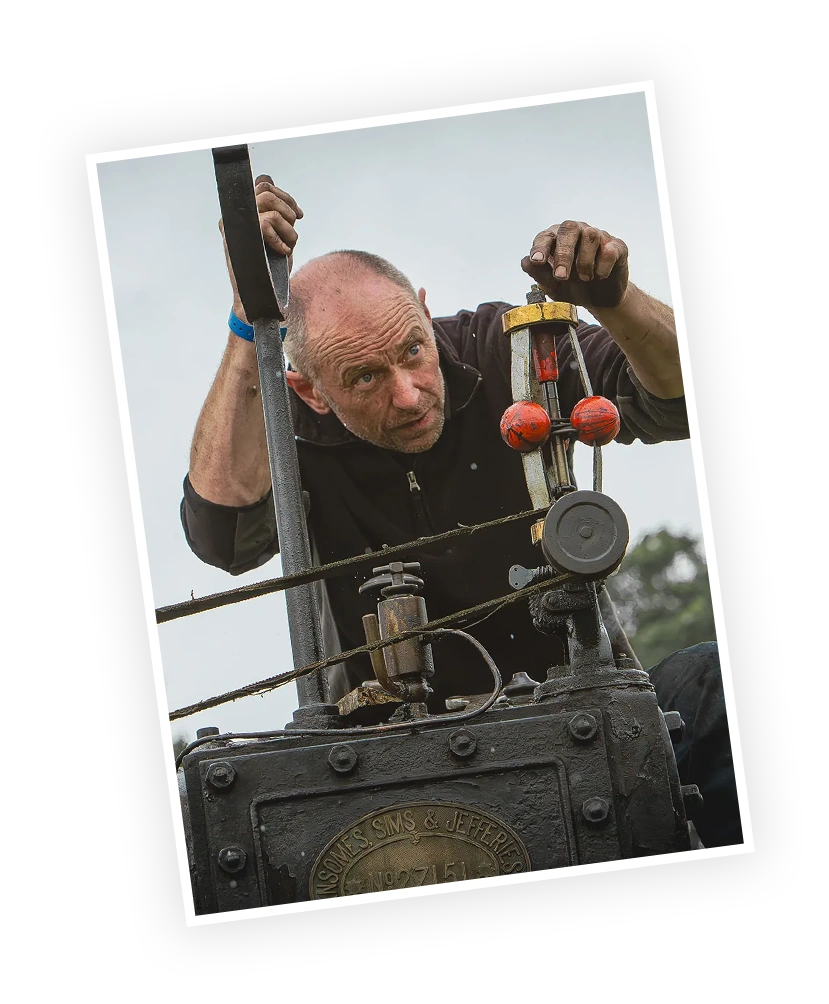 A man inspects the controls of a vintage steam engine, focusing intently on the mechanism. The setting appears to be outdoors, suggesting a heritage or engineering event.