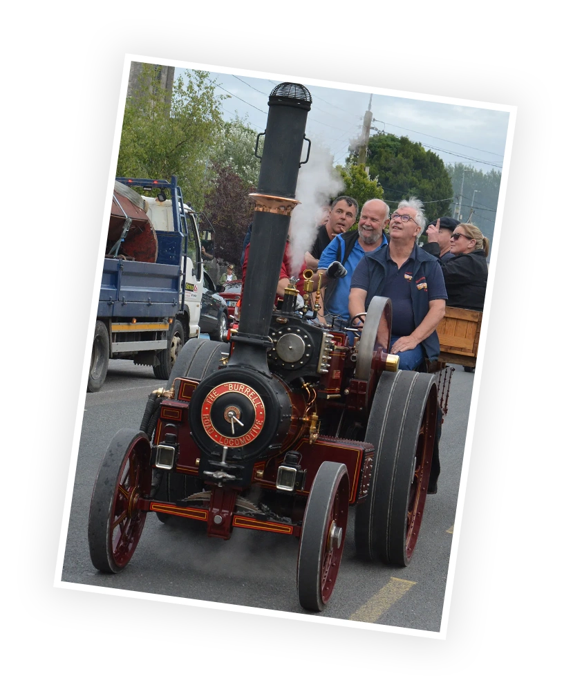Steam-powered traction engine, a Burrell model, is being driven down a road at a local event, with spectators smiling and engaging in the background.