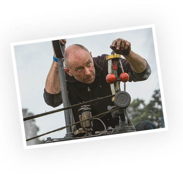 A man inspects the controls of a vintage steam engine, focusing intently on the mechanism. The setting appears to be outdoors, suggesting a heritage or engineering event.