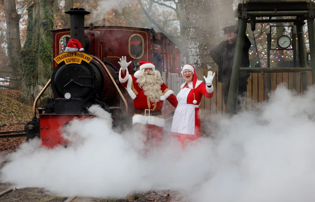 Santa Claus and Mrs Claus pose cheerfully beside the steam locomotive "Stradbally Woodland Express," surrounded by a cloud of steam and holiday decorations, setting a festive atmosphere.