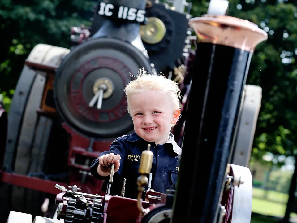 A young boy with blonde hair smiles while sitting at the controls of a vintage steam engine, showcasing enthusiasm for classic machinery in a sunny outdoor setting.
