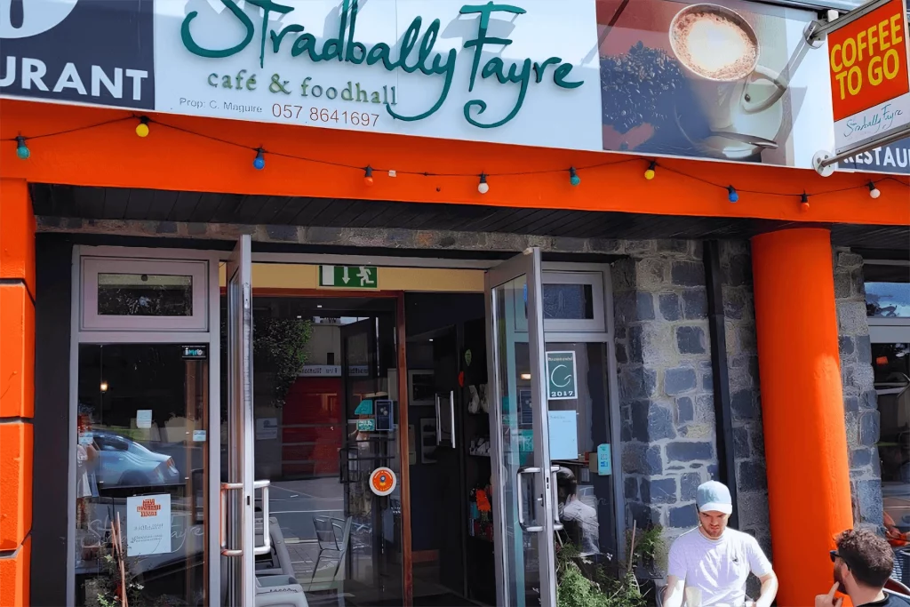 Stradbally Fayre café and foodhall entrance, featuring vibrant orange pillars and decorative lighting, with patrons enjoying the outdoor space. Coffee to go sign visible, highlighting takeaway options.