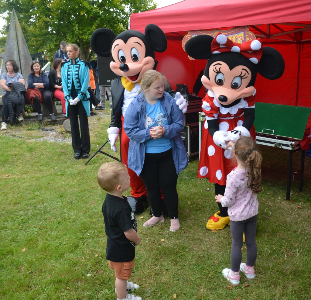 Children interact with mascot characters Mickey and Minnie Mouse at a festive outdoor event, with a small group of onlookers in the background.