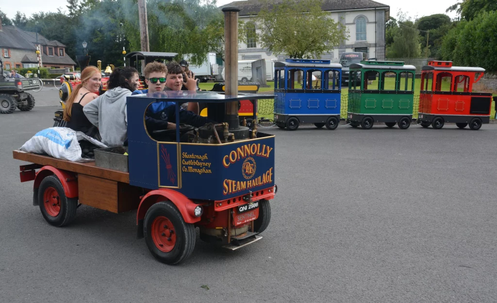 A vintage steam-style vehicle named "Connolly Steam Haulage" with young people aboard, emitting steam, in a festive outdoor setting. Brightly coloured miniature trains are parked in the background, adding to the lively atmosphere.