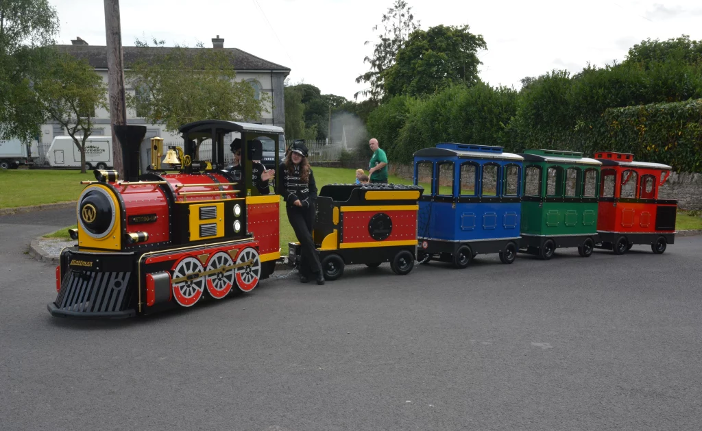 A brightly coloured miniature steam train with a red and yellow engine and several carriages in different colours, stationed on a road surrounded by greenery. Two staff members engage with the camera.