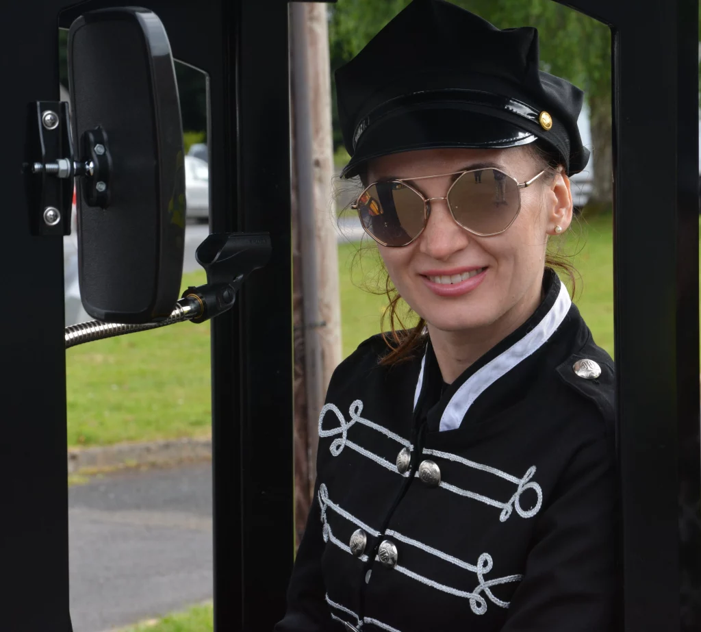 Person wearing a vintage military-style jacket and a hat with sunglasses, seated in a vehicle, smiling at the camera against a green park backdrop.