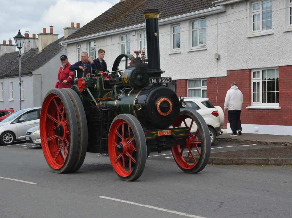 Historic steam tractor driving along a street, with three passengers on board, showcasing vintage machinery alongside residential buildings in an Irish town.