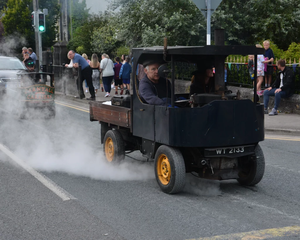 Vintage steam-powered vehicle on a busy street, emitting smoke and surrounded by pedestrians. A man is driving while another small vehicle appears in the background, contributing to the lively scene.