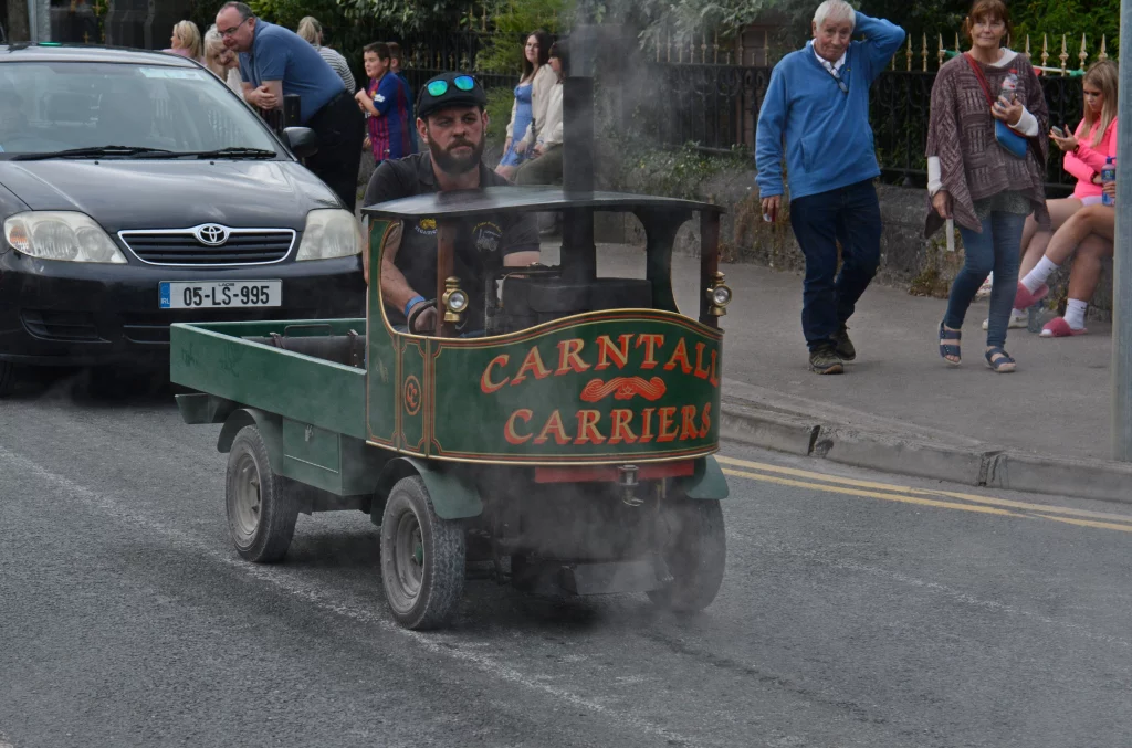 A vintage miniature steam vehicle, branded 'Carntall Carriers', is driving down a street surrounded by pedestrians, with smoke billowing from the engine.