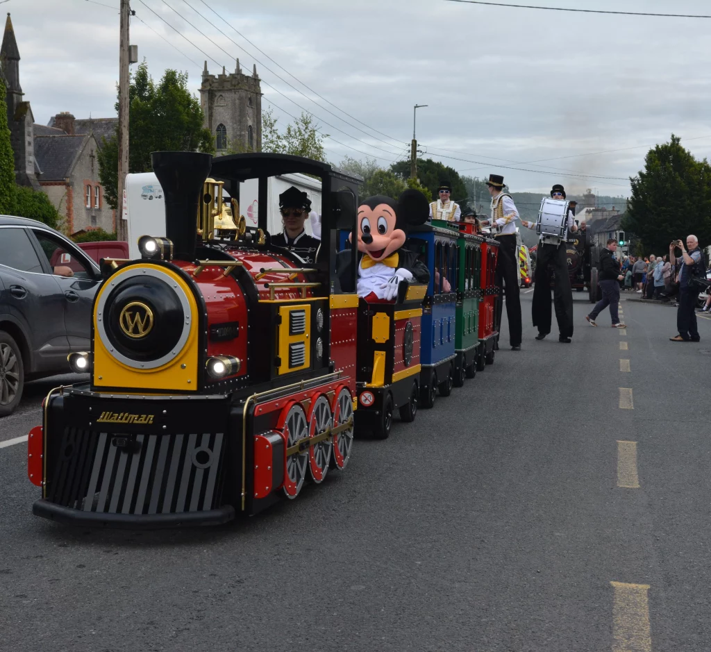 A vibrant train parade featuring a costumed character resembling a mouse, with performers in festive attire and onlookers enjoying the scene, set against a rural backdrop.
