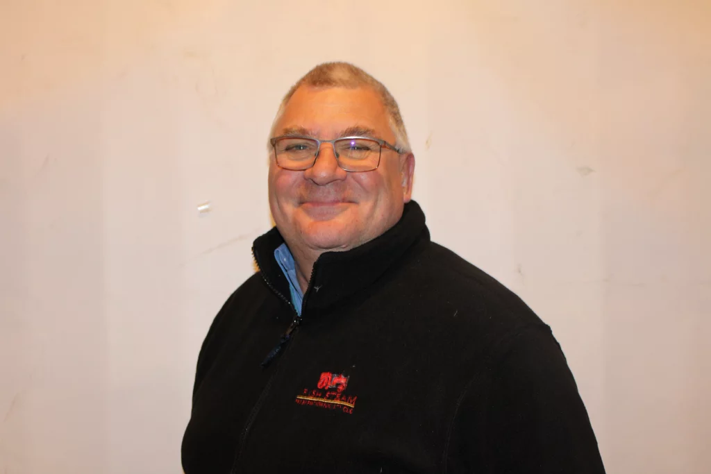 Smiling man wearing spectacles and a black fleece jacket, posing against a plain wall background.