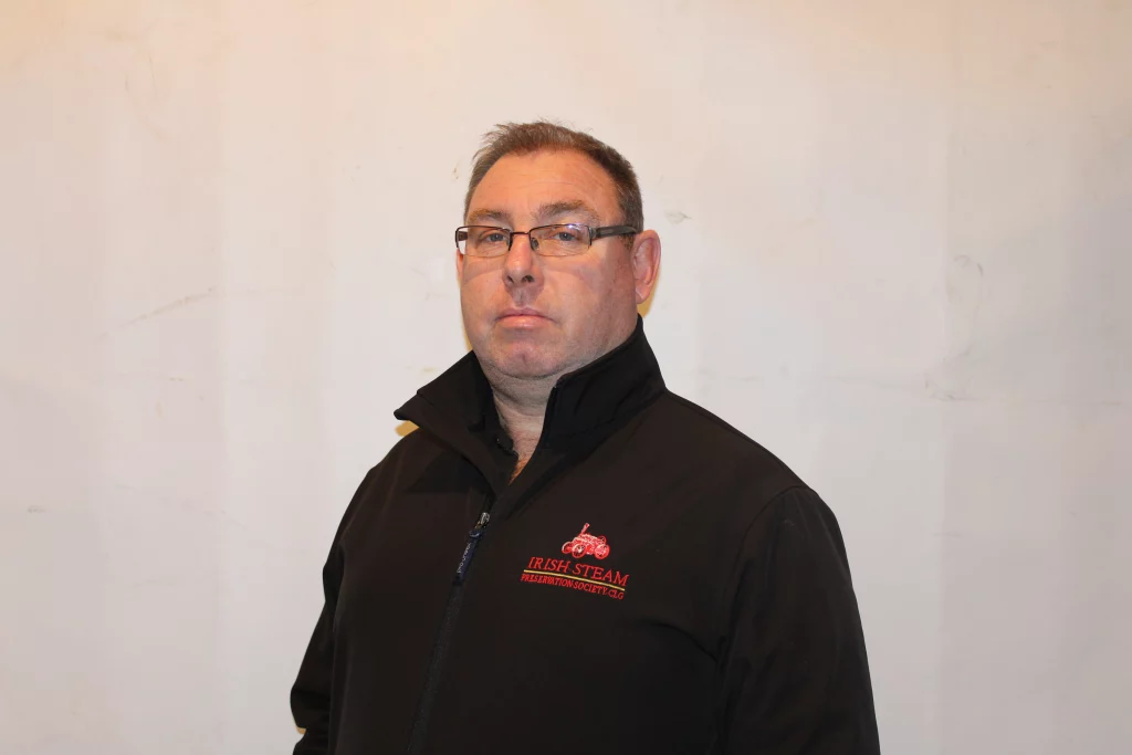 Man in a black zip-up jacket with the "Irish Steam Preservation Society" logo, standing in front of a plain white wall.