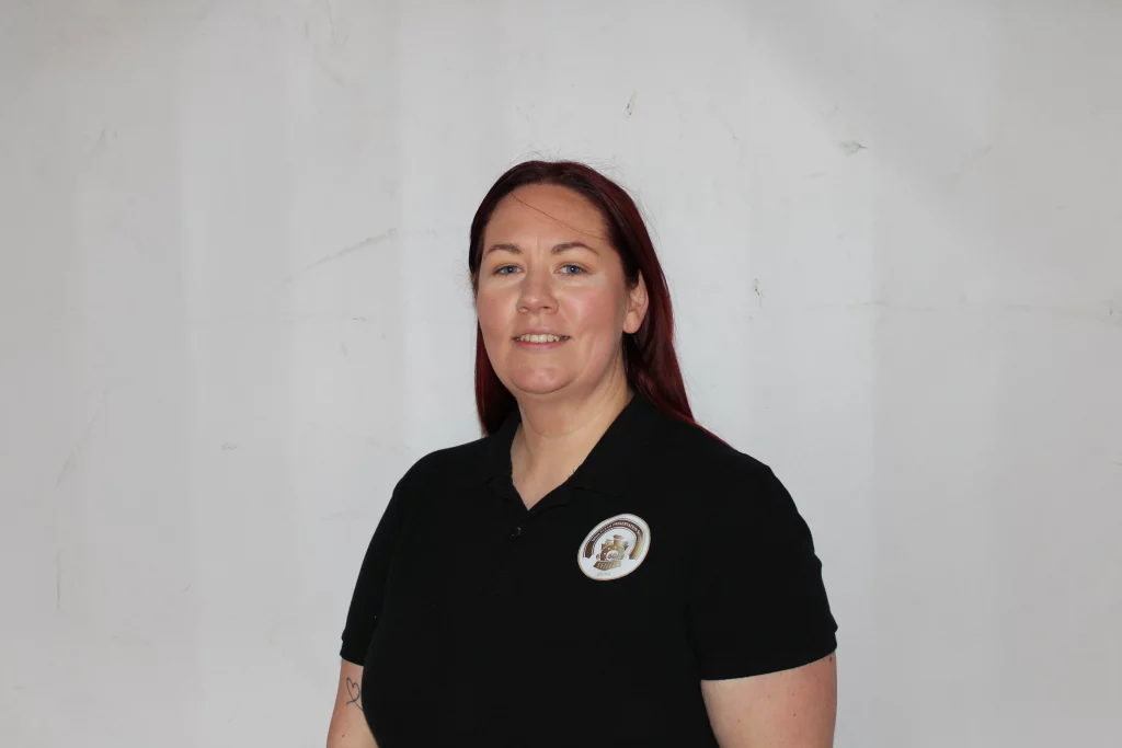 Woman with red hair smiles confidently, wearing a black polo shirt with a badge, set against a plain white background.