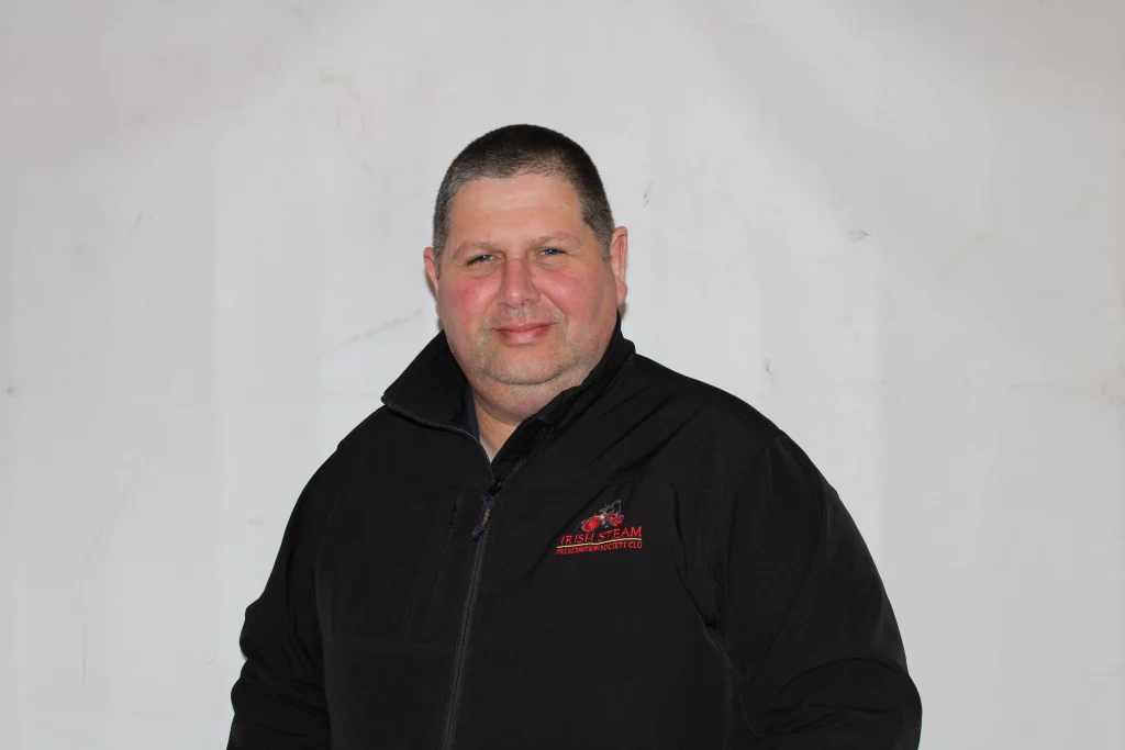 Man smiling in a black fleece jacket with a logo, standing against a plain white background.