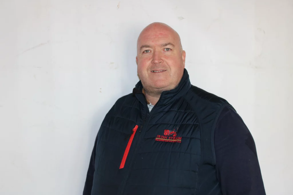 Man wearing a navy padded vest with a red zip and embroidered logo, smiling against a plain white background.