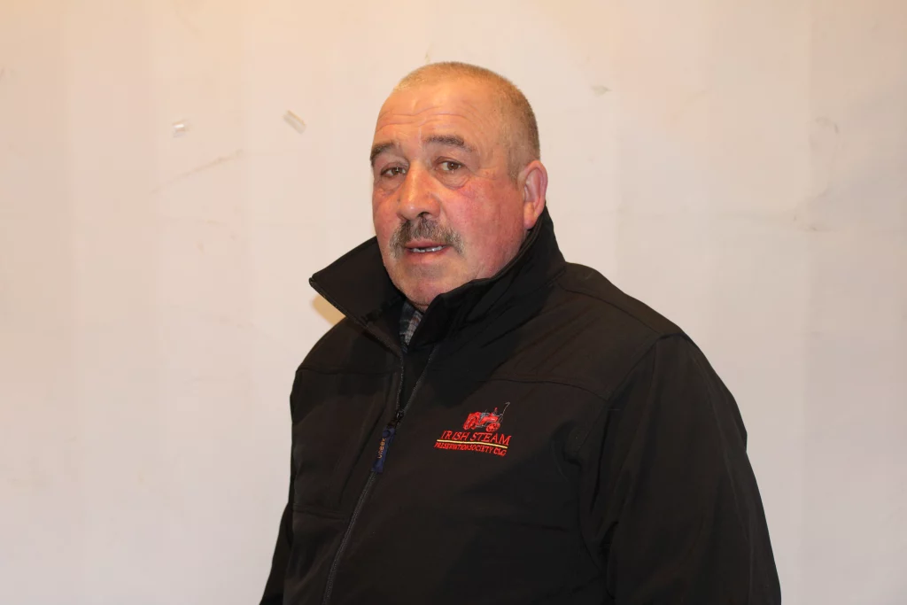 A man wearing a black jacket with a red logo, standing against a plain white backdrop.