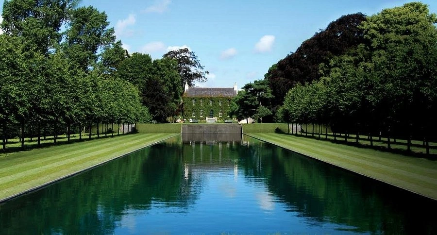 Reflective canal flanked by manicured lawns and tree-lined paths at Ballintubbert Gardens, with the historic ivy-clad house in the distance under a bright blue sky.