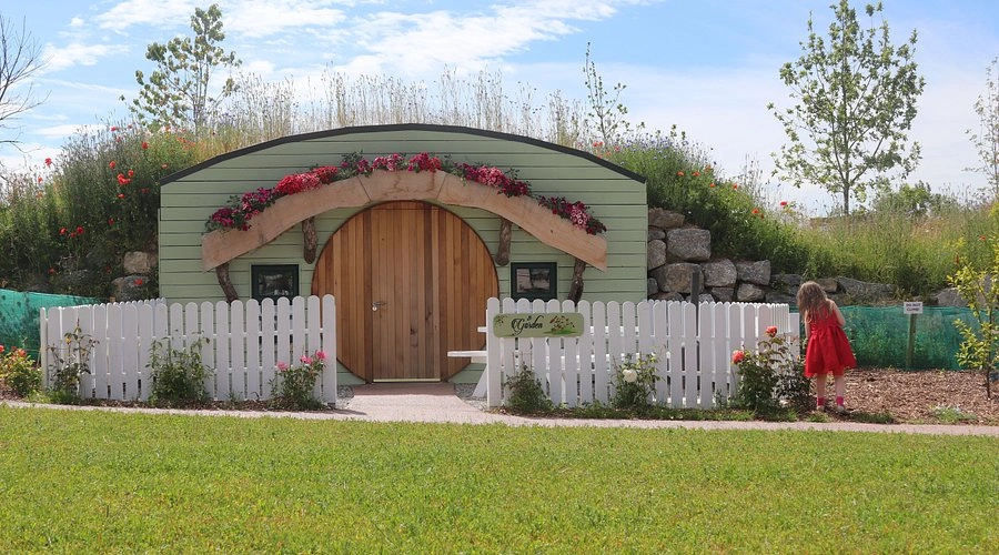 A whimsical hobbit-style glamping with a rounded wooden door, adorned with flowers, surrounded by a white picket fence.