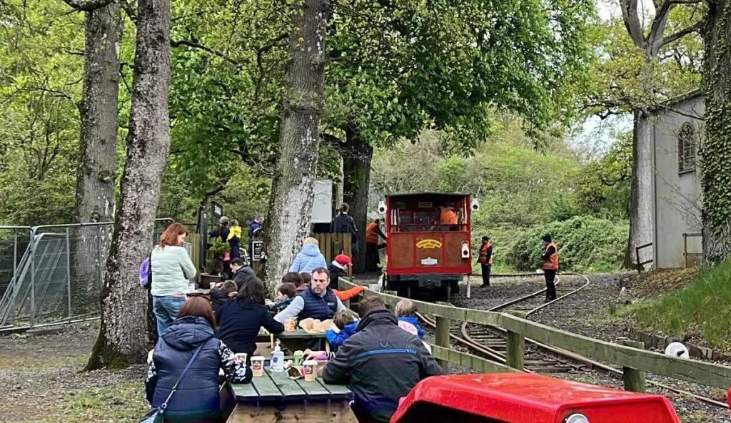 Families enjoying a meal at a picnic table near a vintage train station, featuring a red train and lush greenery in the background.