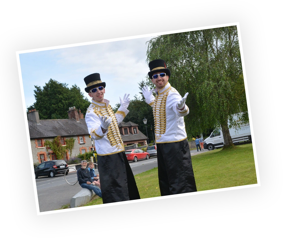 Stilt walkers in stylish costumes pose cheerfully outdoors during a public event, creating a festive atmosphere. In the background, onlookers can be seen enjoying the scene.