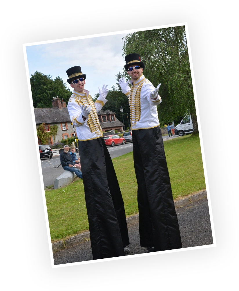 Stilt walkers in stylish costumes pose cheerfully outdoors during a public event, creating a festive atmosphere. In the background, onlookers can be seen enjoying the scene.