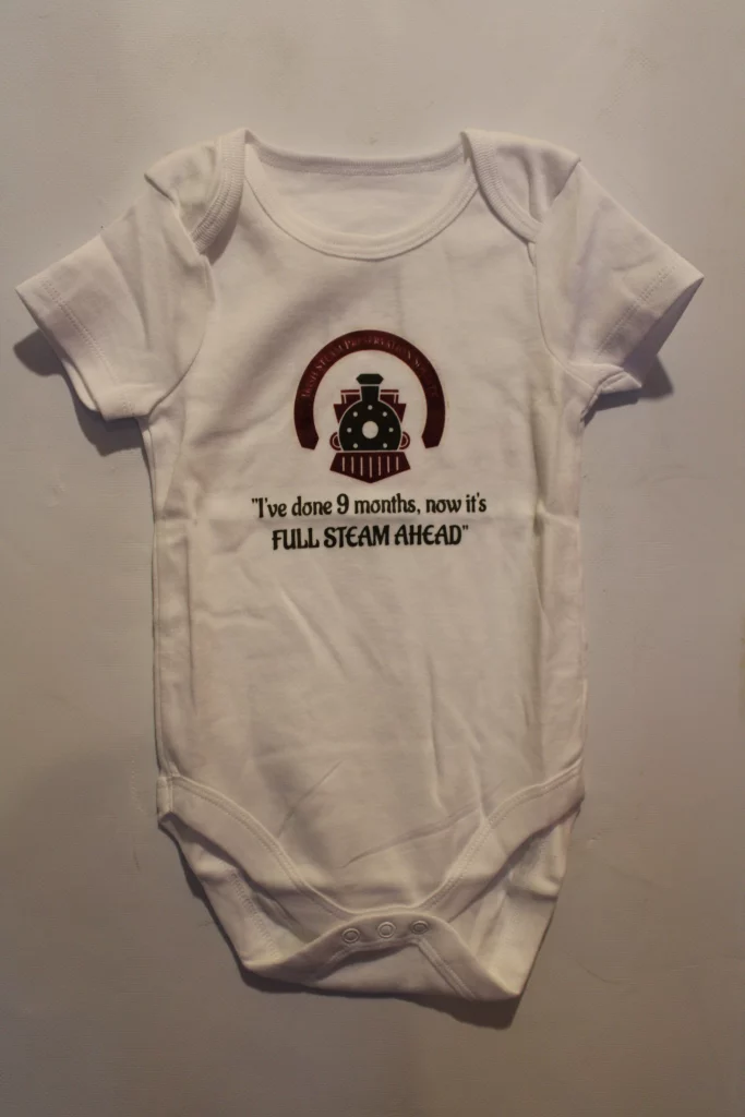 White baby bodysuit featuring the phrase, "I've done 9 months, now it's FULL STEAM AHEAD," alongside a graphic of a steam train, celebrating the journey of pregnancy and the arrival of a new baby.