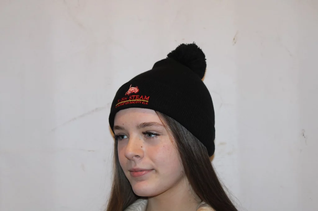 Teen wearing a black bobble hat with a pom-pom, featuring the logo of the Irish Steam Society.
