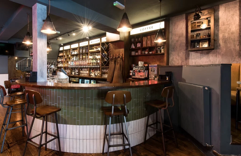 Stylish bar interior featuring a curved wooden counter, high stools, and an extensive selection of spirits displayed on shelves. Warm lighting creates an inviting atmosphere, ideal for socialising. Coffee station is visible in the background.