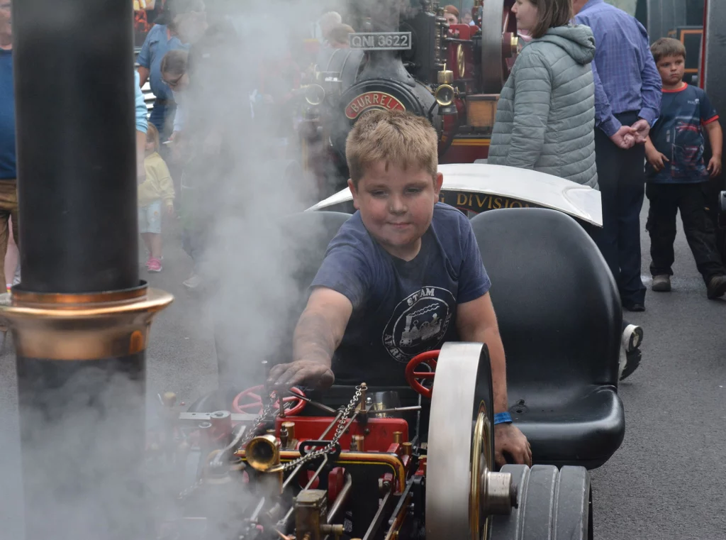 Young boy interacting with a miniature steam engine, surrounded by spectators. The scene is filled with steam, highlighting the vintage machinery and the boy's enthusiasm.