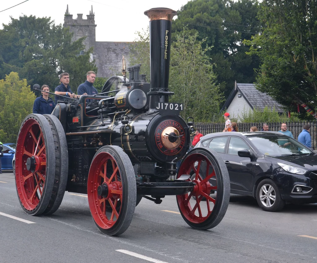 A vintage steam engine, marked "Foster," with red wheels, leads a parade.