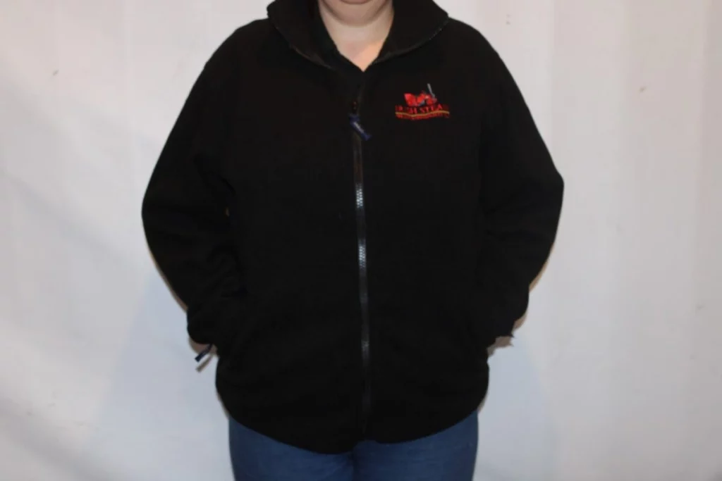 Black fleece jacket with a red embroidered logo, worn by a person with hands in pockets, standing against a plain background.