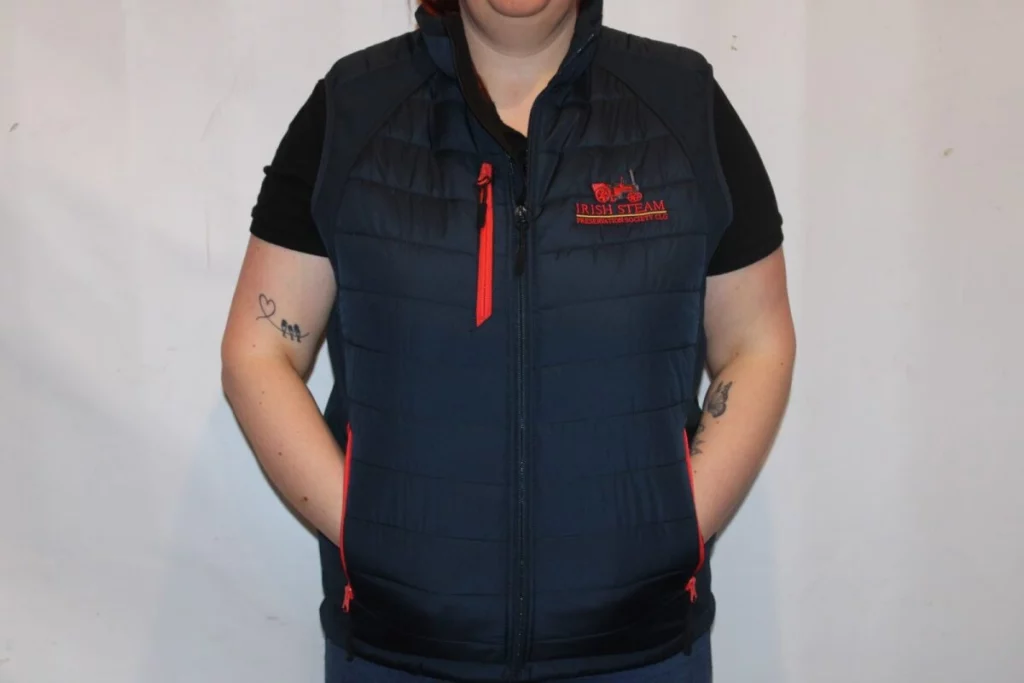 A woman wearing a navy padded gilet with red accents, featuring the logo of the Irish Steam Preservation Society, stands with her hands in her pockets against a plain background.