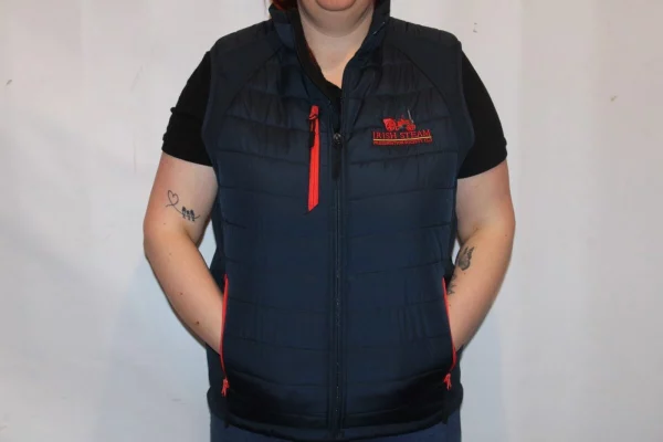 A woman wearing a navy padded gilet with red accents, featuring the logo of the Irish Steam Preservation Society, stands with her hands in her pockets against a plain background.