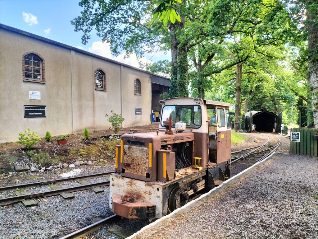 Rusty locomotive parked beside a railway track, with a backdrop of trees and a building featuring signs for Stanground Railway Tours. The scene conveys a historical railway setting, inviting visitors to explore the site.