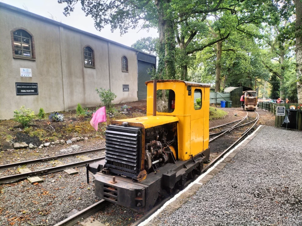 Yellow industrial locomotive parked beside a gravel track at Stradbally Woodland Railway, with trees and a building in the background, showcasing a vintage rail experience.