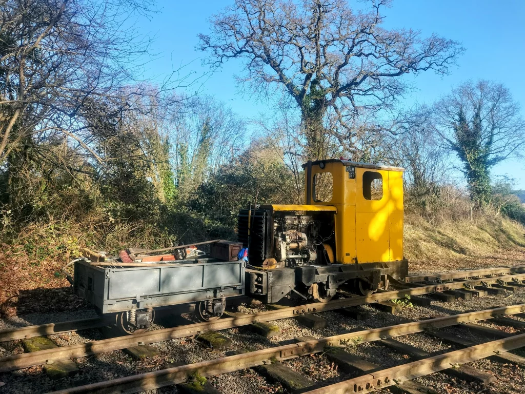 Vintage yellow diesel locomotive parked beside a rustic railway track, accompanied by a grey flatbed wagon loaded with tools, surrounded by trees under a clear blue sky.