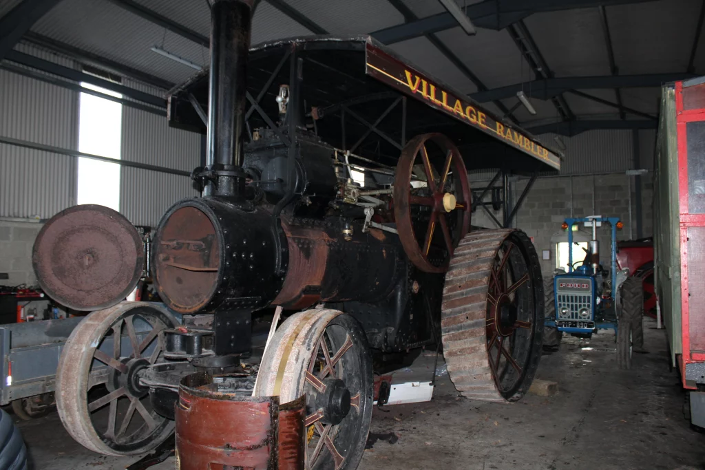 Steam traction engine "Village Rambler" displayed in a shed, showcasing its large wheels and historic design, alongside a blue tractor in the background.