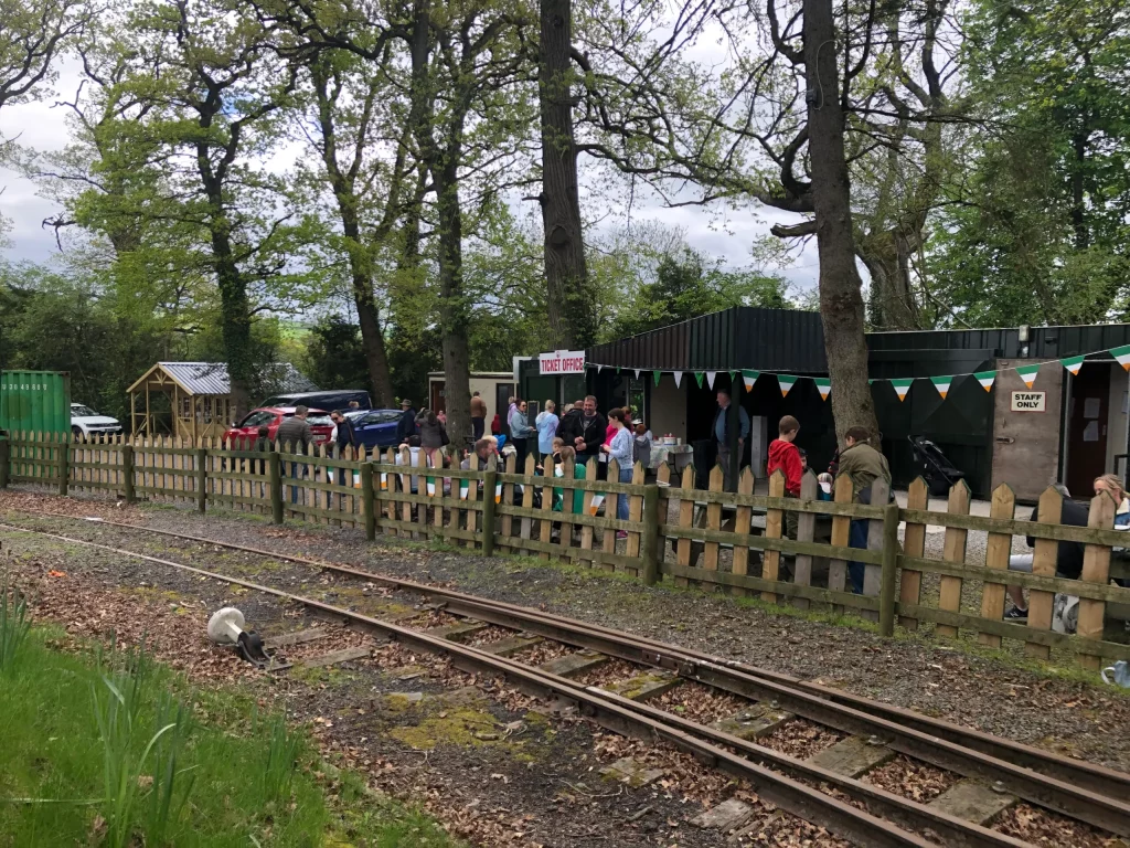 Queue of visitors outside a ticket office at a scenic railway station, surrounded by greenery and wooden fencing, with cars parked nearby. Bunting hangs across the office, suggesting a festive atmosphere.