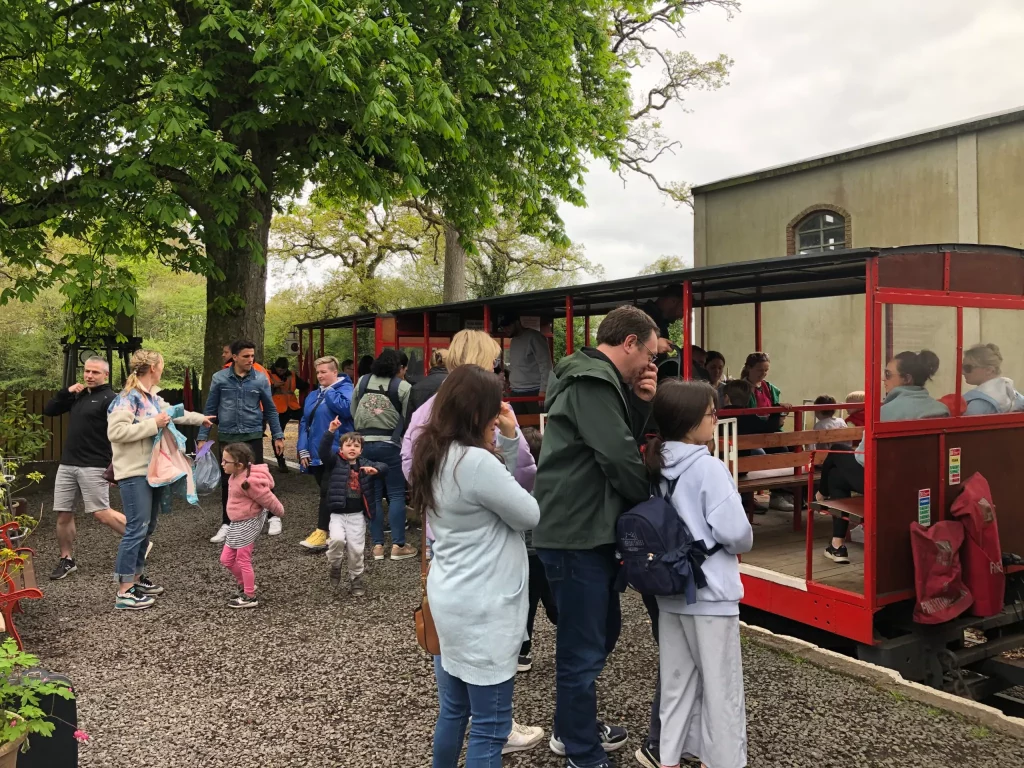 Families and children gather at a small train station, waiting for a ride on a vintage train surrounded by greenery. The atmosphere is lively, with people chatting and enjoying their day.