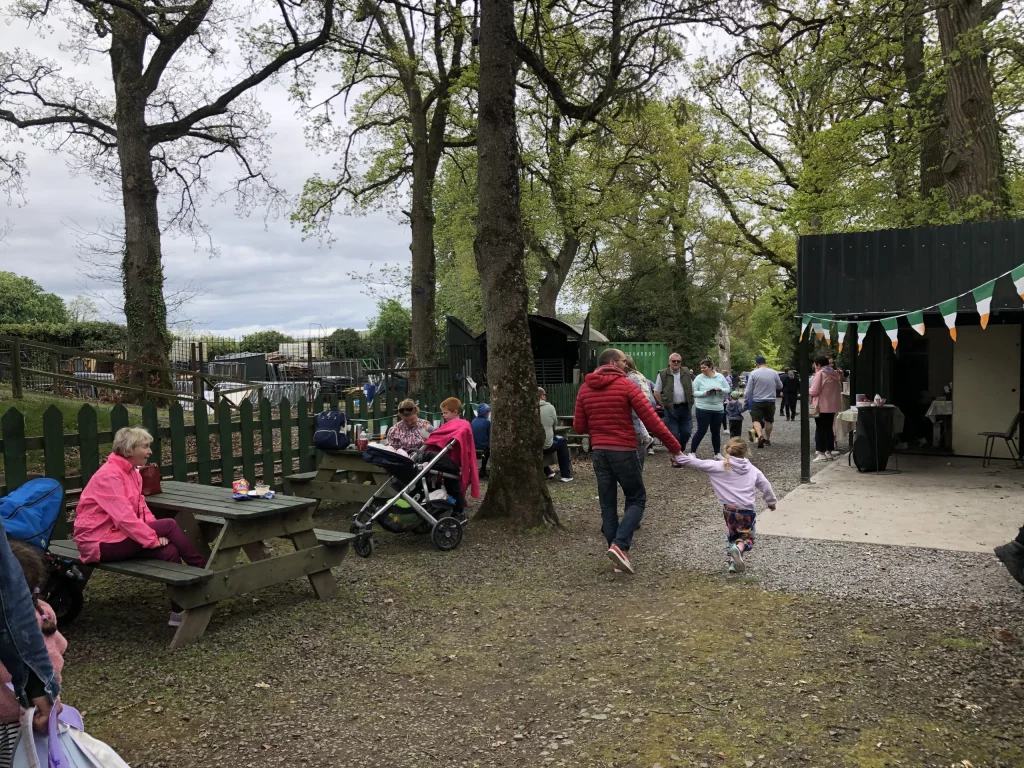 Families enjoy a lively outdoor gathering, with children playing and adults socialising near picnic tables and a food stall, surrounded by trees and greenery.