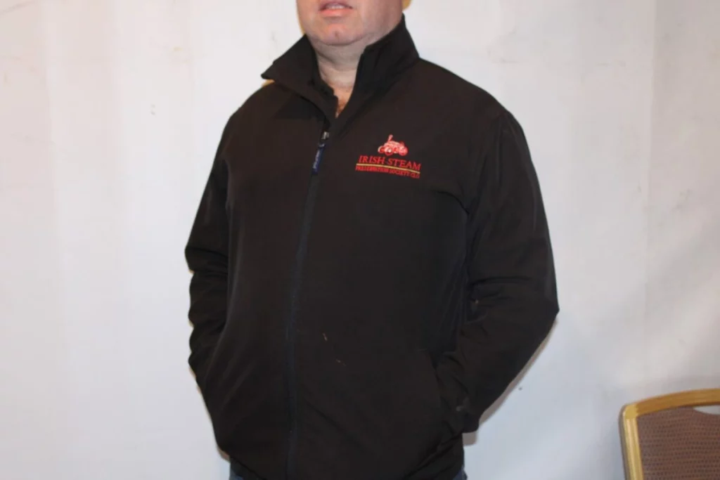 A man wearing a black zip-up jacket featuring the "Irish Steam Preservation Society" logo stands against a plain backdrop.