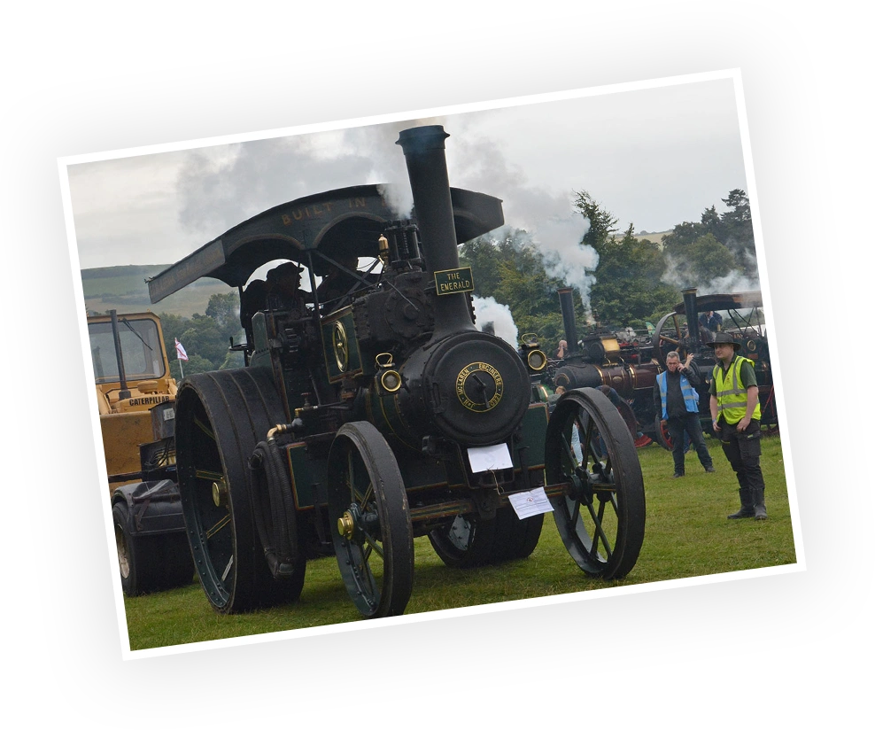 Historic steam engine "The Emerald" on display at an outdoor event, surrounded by spectators and emitting steam. The vehicle showcases vintage engineering with a clearly visible brass chassis and intricately designed wheels.