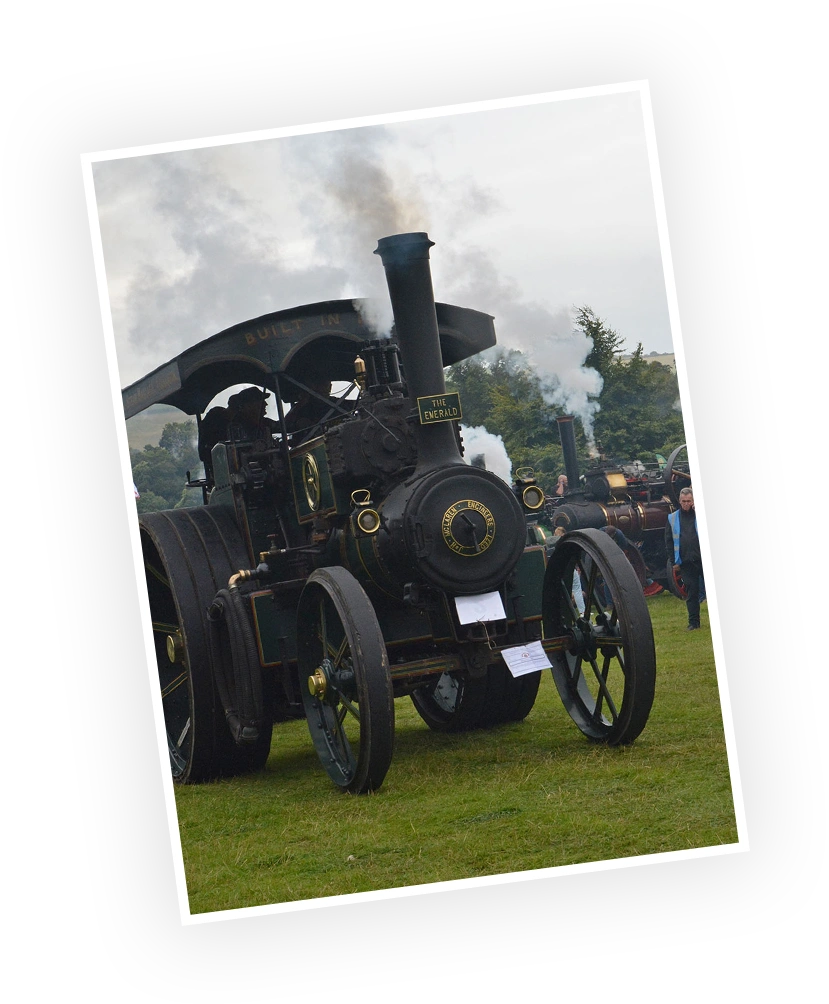 Historic steam engine "The Emerald" on display at an outdoor event, surrounded by spectators and emitting steam. The vehicle showcases vintage engineering with a clearly visible brass chassis and intricately designed wheels.