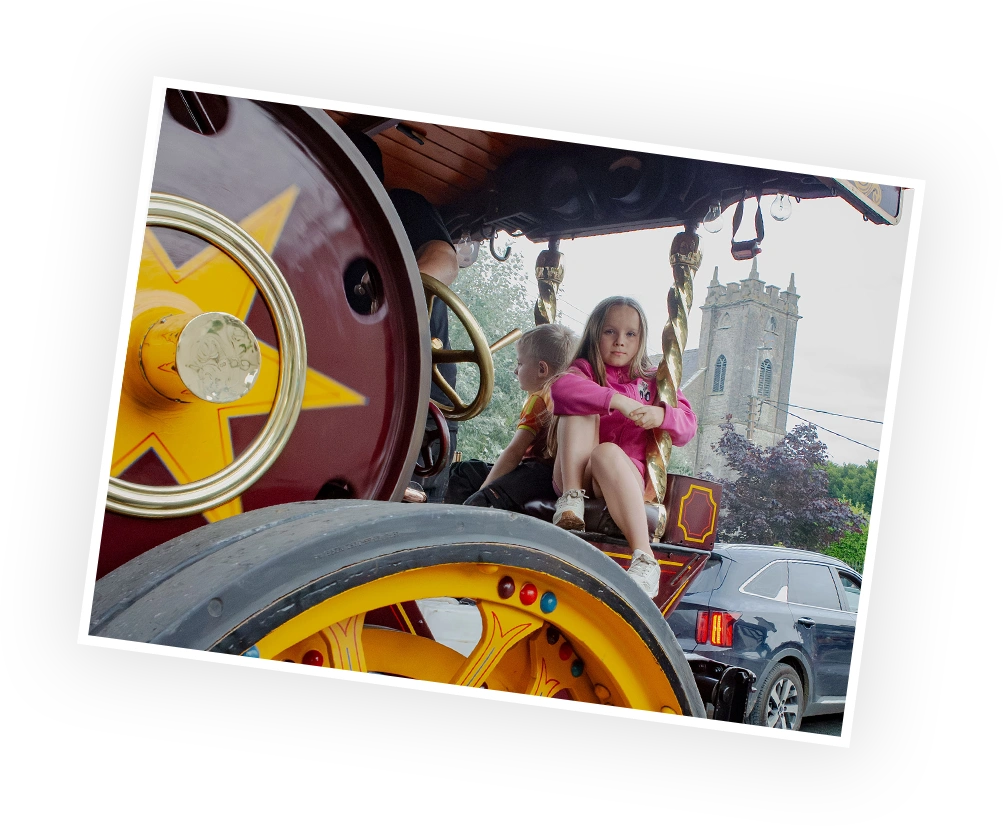A young girl sits playfully on a brightly painted, oversized wheel of a vintage steam engine, with a historical building visible in the background. The vibrant colours and whimsical design highlight the charm of a festive event.