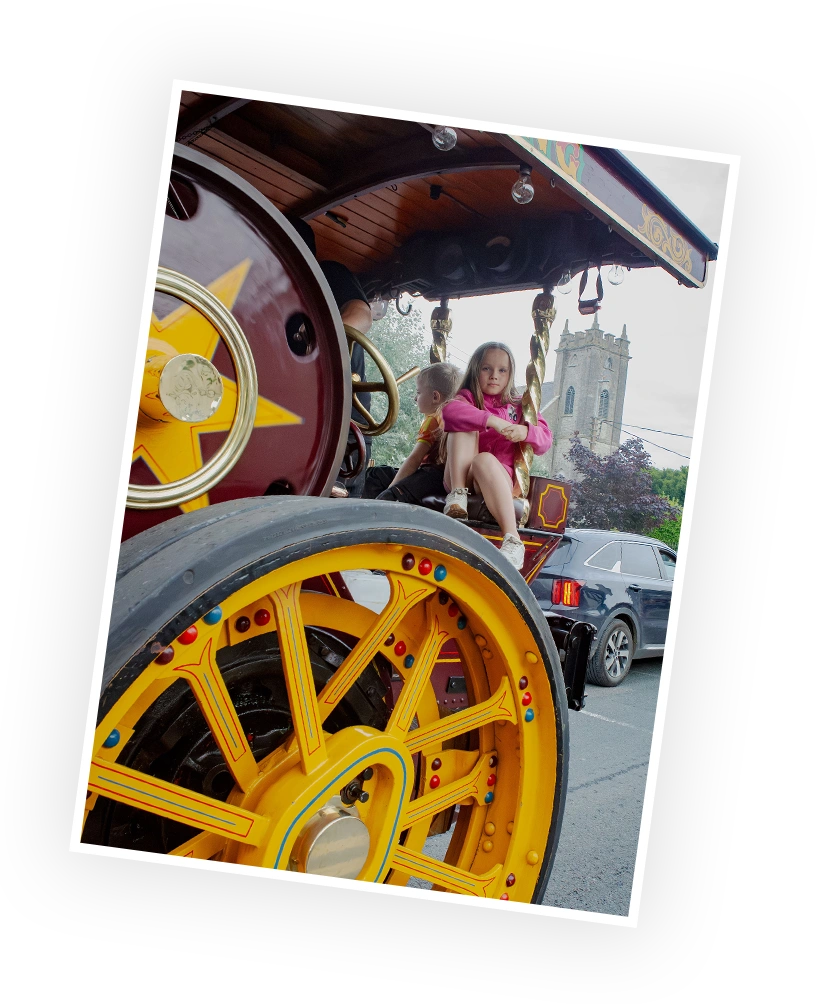 A young girl sits playfully on a brightly painted, oversized wheel of a vintage steam engine, with a historical building visible in the background. The vibrant colours and whimsical design highlight the charm of a festive event.