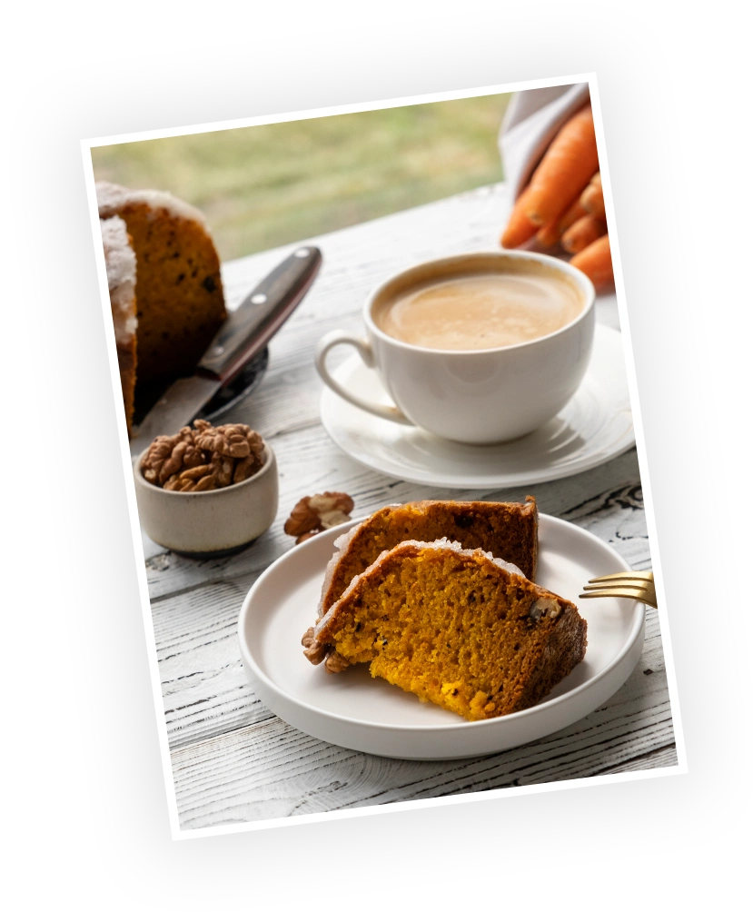 Warm carrot cake slices beside a cup of coffee, with a bowl of walnuts in the foreground, set against a rustic wooden table.