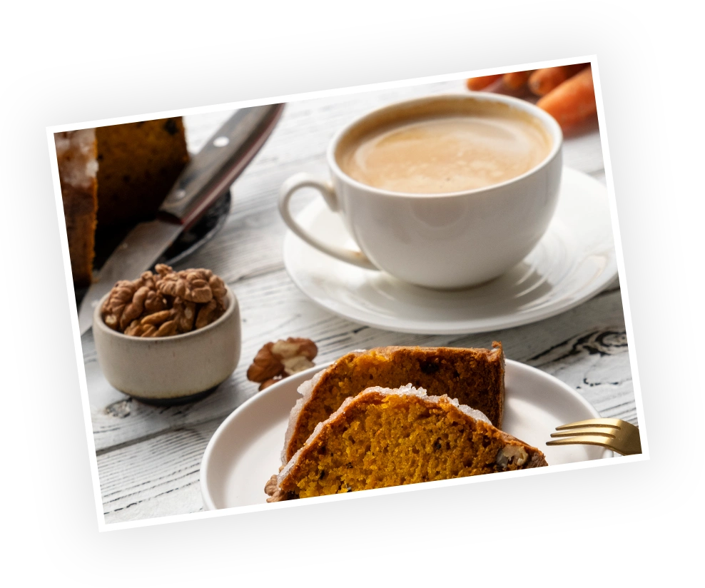 Warm carrot cake slices beside a cup of coffee, with a bowl of walnuts in the foreground, set against a rustic wooden table.
