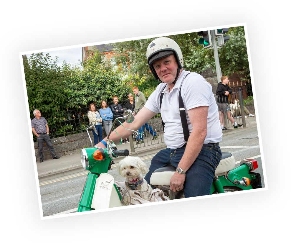 A man wearing a helmet sits on a vintage scooter, holding the handlebars, with a small dog beside him. Pedestrians watch in the background, surrounded by greenery.
