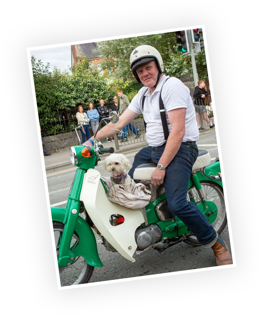 A man wearing a helmet sits on a vintage scooter, holding the handlebars, with a small dog beside him. Pedestrians watch in the background, surrounded by greenery.