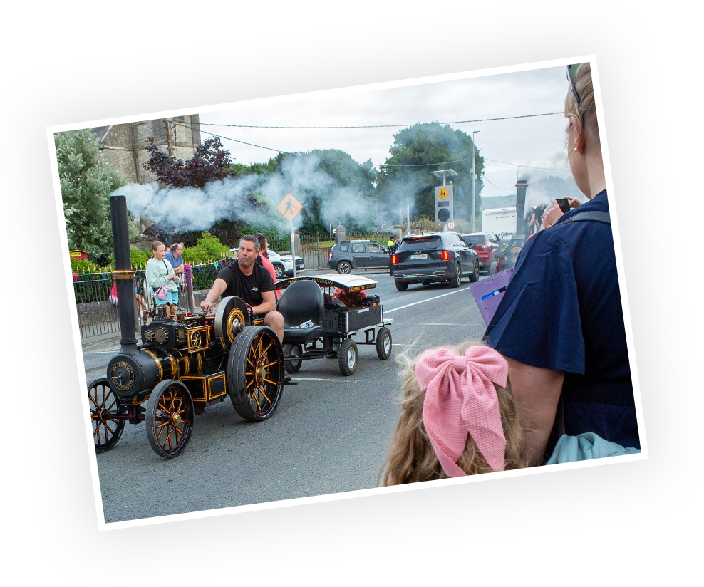 A vintage steam engine parades along a street during a local event, with a man operating the vehicle and children watching in excitement.